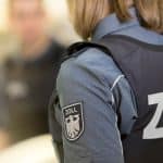 Ein Zollbeamter in Uniform mit dem Wappen des Zolls auf dem Ärmel, im Hintergrund unscharf eine Person und ein Klemmbrett sichtbar.