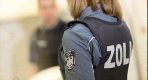 Ein Zollbeamter in Uniform mit dem Wappen des Zolls auf dem Ärmel, im Hintergrund unscharf eine Person und ein Klemmbrett sichtbar.