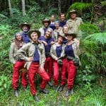 Gruppenfoto der Dschungelcamp 2025-Teilnehmer in ihrer typischen Camp-Bekleidung bestehend aus beigen Jacken, roten Hosen und Hüten, umgeben von tropischer Vegetation.