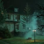 Das Nolden-Haus bei Nacht, umgeben von Nebelschwaden und schattigen Bäumen. Die unheimliche Beleuchtung und der dichte Rauch verstärken die mysteriöse Atmosphäre, die das Gebäude umgibt. Ein einzelner Laternenpfahl wirft ein warmes Licht auf den Garten, während das Haus selbst dunkel und geheimnisvoll erscheint.