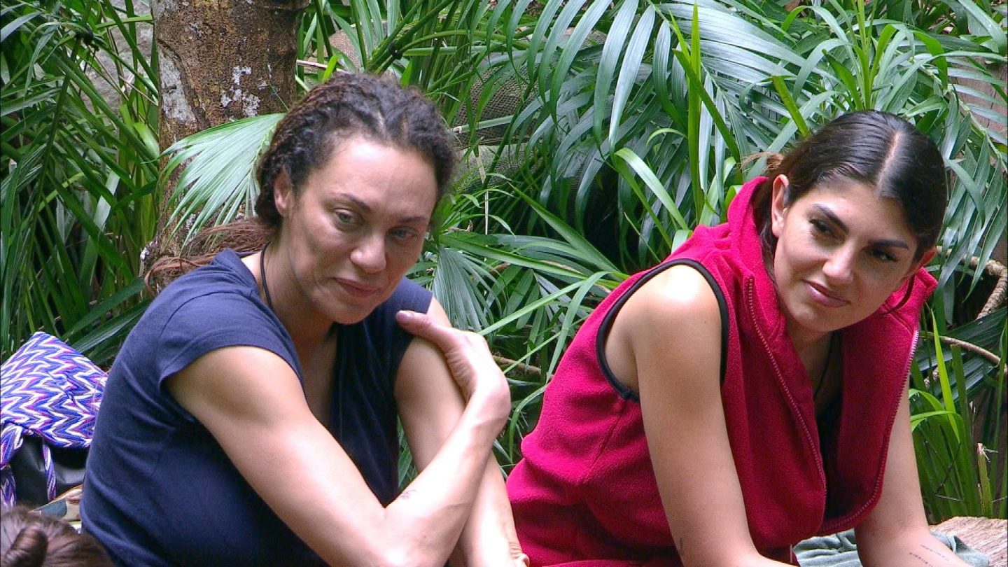 Zwei Frauen sitzen im Dschungelcamp inmitten grüner Pflanzen. Links ist Lilly Becker in einem dunkelblauen T-Shirt zu sehen, die nachdenklich wirkt und ihren Arm auf dem Knie abstützt. Rechts sitzt Yeliz Koc in einer roten Weste, sie schaut mit einem skeptischen Gesichtsausdruck zur Seite. Im Gespräch äußert Lilly ihre Meinung über Mitcamper Sam Dylan, während Yeliz anderer Meinung zu sein scheint. Der Dschungel mit seinen dichten Pflanzen bildet den Hintergrund