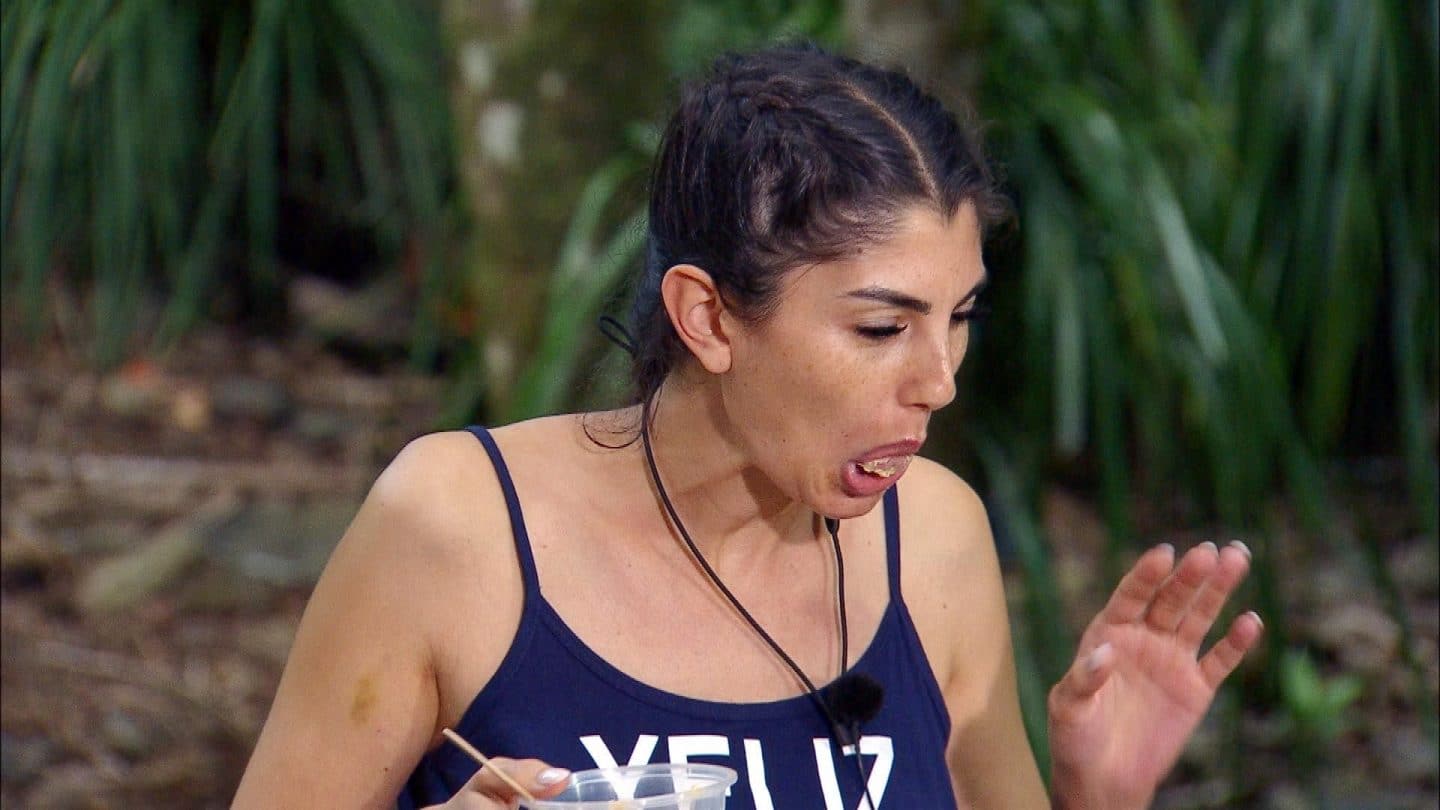 Yeliz Koc während der Dschungelprüfung „Dschungel-Kiosk“. Mit verzogenem Gesicht kämpft sie gegen den Ekel an, während sie eine Schale mit einer unappetitlichen Mahlzeit in der Hand hält. Der Dschungelhintergrund ist mit grüner Vegetation umgeben.