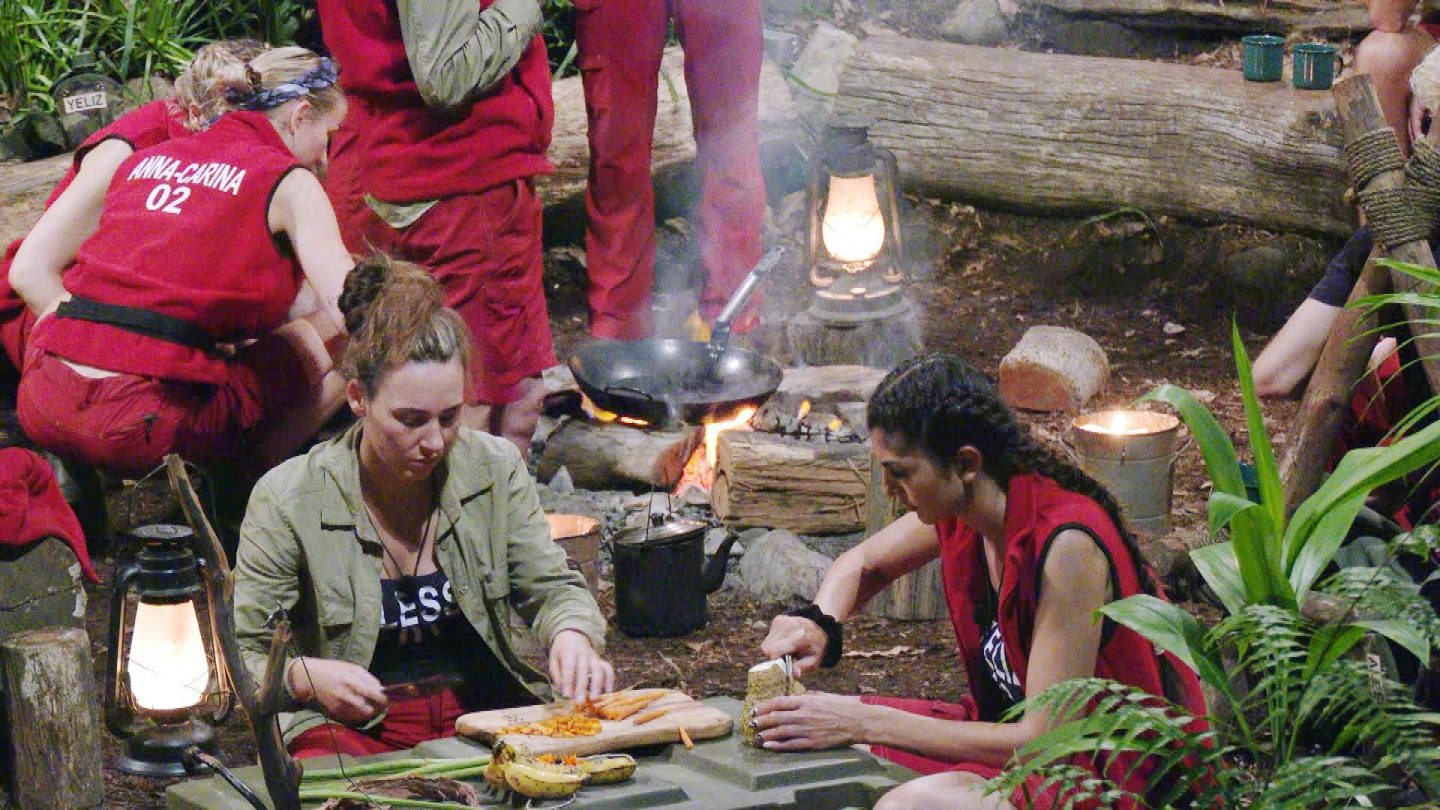 Dschungelcamp 2025: Alessia Herren (links) und Yeliz Koc (rechts) schneiden gemeinsam Gemüse für die Camp-Mahlzeit, während im Hintergrund das Feuer lodert und andere Camper sich um die Zubereitung kümmern.