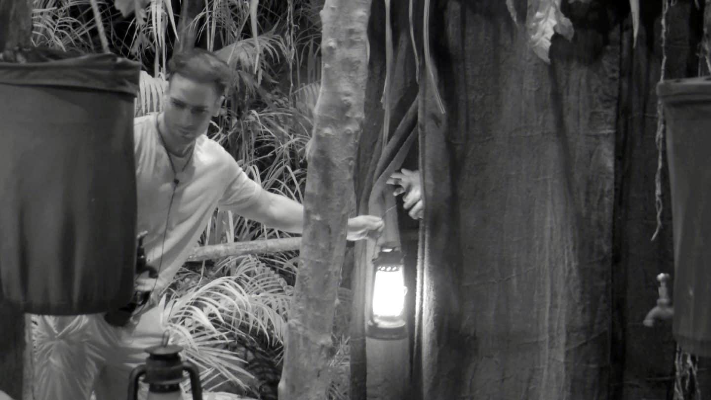 Eine nächtliche Szene im Dschungelcamp in Schwarz-Weiß. Maurice Dziwak steht vor einer provisorischen Toilettenanlage aus Stoffvorhängen. Er hält eine leuchtende Laterne in der Hand und reicht sie einer Person, deren Hand aus dem Vorhang herausragt. Maurice trägt ein helles Shirt und eine Kette um den Hals. Die Umgebung ist von dichter Dschungelvegetation und Holzkonstruktionen geprägt, die das Camp einrahmen.