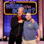 Tom Gerhardt und Axel Stein stehen gemeinsam auf der Bühne der TV-Quizshow "Wer weiß denn sowas?" vor dem großen, beleuchteten Logo der Sendung. Tom Gerhardt trägt eine schwarze Strickjacke über einem roten Shirt und zeigt mit dem Finger auf Axel Stein, während er ihn freundlich anlächelt. Axel Stein trägt ein blaues T-Shirt mit einem gelben Logo und eine beige Hose, lächelt ebenfalls und hat seinen Arm um die Schulter von Tom Gerhardt gelegt. Die Kulisse zeigt eine Backsteinwand mit stimmungsvoller Beleuchtung.
