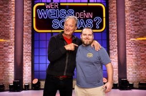 Tom Gerhardt und Axel Stein stehen gemeinsam auf der Bühne der TV-Quizshow "Wer weiß denn sowas?" vor dem großen, beleuchteten Logo der Sendung. Tom Gerhardt trägt eine schwarze Strickjacke über einem roten Shirt und zeigt mit dem Finger auf Axel Stein, während er ihn freundlich anlächelt. Axel Stein trägt ein blaues T-Shirt mit einem gelben Logo und eine beige Hose, lächelt ebenfalls und hat seinen Arm um die Schulter von Tom Gerhardt gelegt. Die Kulisse zeigt eine Backsteinwand mit stimmungsvoller Beleuchtung.