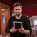 Ralph Gassmann als Andreas hält nachdenklich ein gerahmtes Foto in den Händen, während er in einem roten Zimmer steht, das von Familienbildern und Alltagsgegenständen geprägt ist.