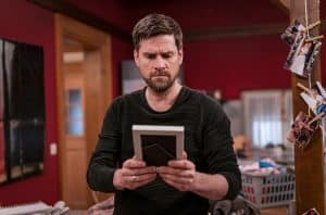 Ralph Gassmann als Andreas hält nachdenklich ein gerahmtes Foto in den Händen, während er in einem roten Zimmer steht, das von Familienbildern und Alltagsgegenständen geprägt ist.