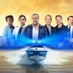 Das Team der Krimiserie „WaPo Duisburg“ vor einer malerischen Kulisse mit Hafen, Containern und Industrieanlagen im Hintergrund. Im Vordergrund ein Polizeiboot mit der Aufschrift „Polizei Duisburg“ auf einem Fluss. Die Hauptfiguren der Serie stehen in Polizeiuniformen und Zivilkleidung nebeneinander, darunter Markus John und weitere Darsteller, die die Ermittler und Teammitglieder verkörpern. Der Himmel ist in warmen Gelb- und Orangetönen gehalten und strahlt eine dramatische Atmosphäre aus.