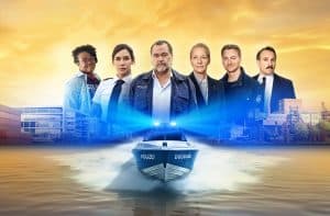 Das Team der Krimiserie „WaPo Duisburg“ vor einer malerischen Kulisse mit Hafen, Containern und Industrieanlagen im Hintergrund. Im Vordergrund ein Polizeiboot mit der Aufschrift „Polizei Duisburg“ auf einem Fluss. Die Hauptfiguren der Serie stehen in Polizeiuniformen und Zivilkleidung nebeneinander, darunter Markus John und weitere Darsteller, die die Ermittler und Teammitglieder verkörpern. Der Himmel ist in warmen Gelb- und Orangetönen gehalten und strahlt eine dramatische Atmosphäre aus.