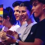 Ein jubelndes Publikum bei einer Veranstaltung, eine Frau hält eine kleine deutsche Flagge in der Hand, während andere applaudieren. Die Szene ist in blauem Bühnenlicht getaucht, was eine festliche Stimmung vermittelt.