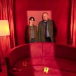 Kommissare Mila Sahin und Klaus Borowski stehen in einem rot ausgeleuchteten Raum vor einem roten Sofa, das mit gelben Beweismarkierungen (8 und 9) versehen ist. Im Hintergrund sind rote Vorhänge und gelbe Lampen zu sehen, die die Szene in eine dramatische, düstere Atmosphäre tauchen.