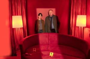 Kommissare Mila Sahin und Klaus Borowski stehen in einem rot ausgeleuchteten Raum vor einem roten Sofa, das mit gelben Beweismarkierungen (8 und 9) versehen ist. Im Hintergrund sind rote Vorhänge und gelbe Lampen zu sehen, die die Szene in eine dramatische, düstere Atmosphäre tauchen.