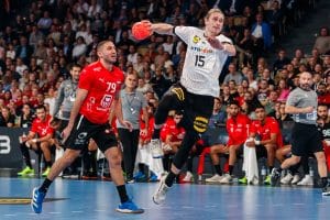Juri Knorr beim Sprungwurf in einem Handballspiel, konzentriert in Aktion, während ein Gegenspieler in rotem Trikot versucht, ihn zu blockieren. Im Hintergrund sind Zuschauer und Teammitglieder zu sehen.