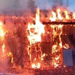Ein Haus, das in voller Ausdehnung brennt, mit hohen Flammen, die aus Fenstern und dem Dach schlagen. Im Vordergrund sind Trümmer und Rauch zu sehen, während im Hintergrund ein Auto und ein weiteres Gebäude teilweise verdeckt sind.