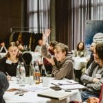 Eine Gruppe von Menschen sitzt in einem Konferenzraum an runden Tischen, die mit Dokumenten, Wasserflaschen und Gläsern bedeckt sind. Im Mittelpunkt hebt eine Person in einem grauen Hoodie motiviert die Hand, während andere Teilnehmer konzentriert Notizen machen oder zuhören. Im Hintergrund sind weitere Personen an Tischen sowie ein Banner mit der Aufschrift „Wir wollen besser“ zu sehen. Die Atmosphäre wirkt engagiert und diskussionsfreudig.