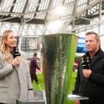 Moderatorin Laura Papendick und RTL-Fußball-Experte Lothar Matthäus stehen im Stadion vor der UEFA Europa League Trophäe und moderieren eine Live-Übertragung. Im Hintergrund sind Fans und das Spielfeld zu sehen