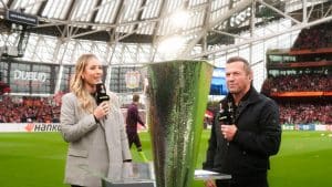 Moderatorin Laura Papendick und RTL-Fußball-Experte Lothar Matthäus stehen im Stadion vor der UEFA Europa League Trophäe und moderieren eine Live-Übertragung. Im Hintergrund sind Fans und das Spielfeld zu sehen