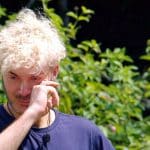 Sam Dylan steht mit traurigem Blick im Dschungelcamp und wischt sich mit der Hand über das Gesicht, als würde er Tränen wegwischen. Er trägt ein dunkelblaues Shirt mit Mikrofon, während im Hintergrund grüne Pflanzen zu sehen sind. Die emotionale Szene deutet auf einen bewegenden Moment im Camp hin.