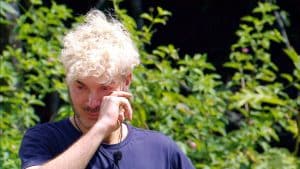 Sam Dylan steht mit traurigem Blick im Dschungelcamp und wischt sich mit der Hand über das Gesicht, als würde er Tränen wegwischen. Er trägt ein dunkelblaues Shirt mit Mikrofon, während im Hintergrund grüne Pflanzen zu sehen sind. Die emotionale Szene deutet auf einen bewegenden Moment im Camp hin.