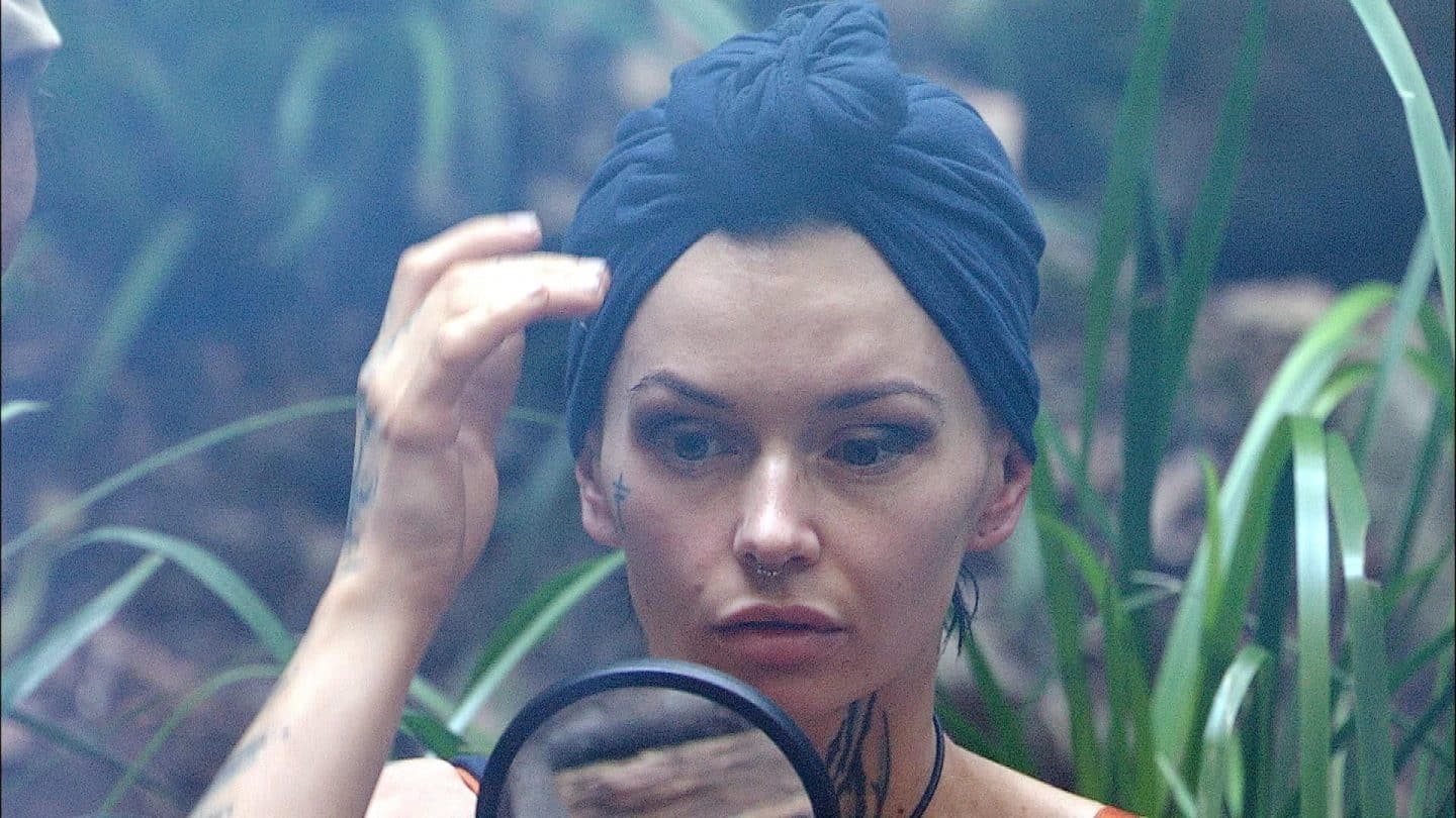 dith Stehfest sitzt im Dschungelcamp und betrachtet ihr Spiegelbild mit ernstem Blick. Mit einem dunklen Turban und markanten Tattoos im Gesicht wirkt sie fokussiert, während grüne Pflanzen im Hintergrund die Szenerie umgeben.