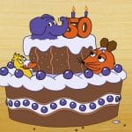 Eine Geburtstagstorte mit Schokoladenverzierung und blauen Beeren. Auf der Torte sitzen die Figuren aus der „Sendung mit der Maus“: der blaue Elefant mit einer "50" in Kerzenform, die orange Maus und die gelbe Ente. Sie feiern gemeinsam das 50-jährige Jubiläum des Elefanten. Im Hintergrund gelb.