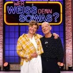 Margarita Broich und Martin Wuttke lächelnd im Studio der Quizshow „Wer weiß denn sowas?“, vor dem leuchtenden Logo der Sendung.