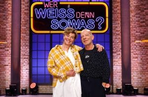 Margarita Broich und Martin Wuttke lächelnd im Studio der Quizshow „Wer weiß denn sowas?“, vor dem leuchtenden Logo der Sendung.