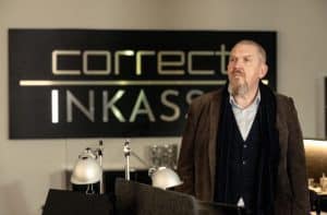 Freddy Schenk (Dietmar Bär) steht nachdenklich im Büro der fiktiven Firma Correct Inkasso. Hinter ihm ist das Firmenschild mit dem Schriftzug „Correct Inkasso“ deutlich zu sehen. Die Szene vermittelt die ernste Atmosphäre der Ermittlungen im Umfeld des umstrittenen Inkasso-Unternehmens.