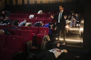 Ein Mann in dunkler Kleidung steht mit erhobenen Händen auf den Stufen eines Kinosaals. Um ihn herum sitzen verängstigte Menschen auf roten Kinositzen, einige halten sich die Hände vors Gesicht. Im Vordergrund liegt eine blutüberströmte Person am Boden, neben ihr eine Waffe. Die Szene ist dramatisch beleuchtet, wodurch die Anspannung und Angst der Anwesenden verstärkt werden.