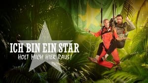 Teaserbild für die RTL-Show "Ich bin ein Star – Holt mich hier raus!". Im Vordergrund schwingen zwei Personen in roten Dschungelanzügen, befestigt an Seilen, durch die tropische Vegetation. Im Hintergrund sind Palmenblätter und ein großes, leicht verschwommenes Banner in den Farben Grün und Gelb zu sehen. Der Schriftzug der Show mit einem Stern als Designelement steht prominent links im Bild.