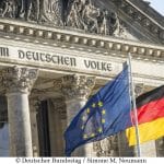 Ein Bild des Reichstagsgebäudes in Berlin, mit den Inschriften „DEM DEUTSCHEN VOLKE“ über dem Eingang. Vor dem Gebäude wehen die deutsche Nationalflagge und die Europaflagge, die zusammen das politische und historische Erbe sowie die europäische Verbundenheit Deutschlands symbolisieren. Die Aufnahme zeigt die Architektur und die Symbole in klaren Details bei Tageslicht.