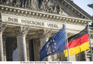 Ein Bild des Reichstagsgebäudes in Berlin, mit den Inschriften „DEM DEUTSCHEN VOLKE“ über dem Eingang. Vor dem Gebäude wehen die deutsche Nationalflagge und die Europaflagge, die zusammen das politische und historische Erbe sowie die europäische Verbundenheit Deutschlands symbolisieren. Die Aufnahme zeigt die Architektur und die Symbole in klaren Details bei Tageslicht.