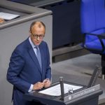 Friedrich Merz im blauen Anzug, vermutlich ein Politiker, steht an einem Rednerpult im Deutschen Bundestag. Er hält ein Dokument in der Hand und spricht vor dem Plenum. Im Hintergrund sind blaue Sitze und eine weitere Person zu sehen.