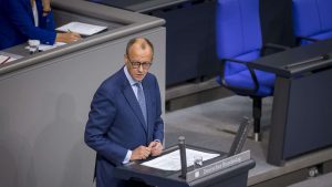 Friedrich Merz im blauen Anzug, vermutlich ein Politiker, steht an einem Rednerpult im Deutschen Bundestag. Er hält ein Dokument in der Hand und spricht vor dem Plenum. Im Hintergrund sind blaue Sitze und eine weitere Person zu sehen.