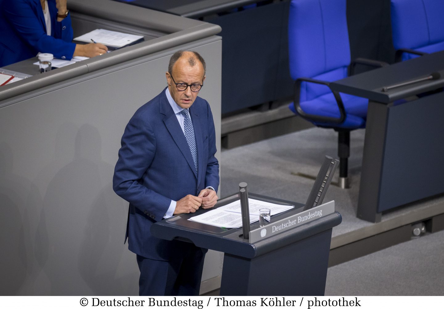Friedrich Merz im blauen Anzug, vermutlich ein Politiker, steht an einem Rednerpult im Deutschen Bundestag. Er hält ein Dokument in der Hand und spricht vor dem Plenum. Im Hintergrund sind blaue Sitze und eine weitere Person zu sehen.