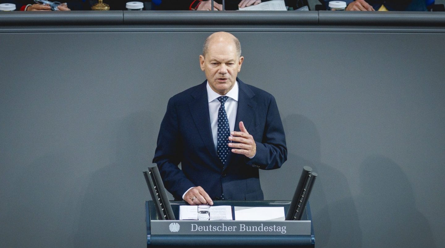 Bundeskanzler Olaf Scholz spricht im Deutschen Bundestag an einem Rednerpult. Er trägt einen dunklen Anzug mit weißem Hemd und blauer Krawatte. Im Hintergrund sind andere Abgeordnete zu sehen.