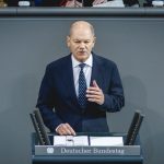 Bundeskanzler Olaf Scholz spricht im Deutschen Bundestag an einem Rednerpult. Er trägt einen dunklen Anzug mit weißem Hemd und blauer Krawatte. Im Hintergrund sind andere Abgeordnete zu sehen.