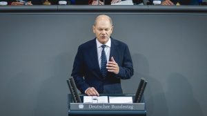 Bundeskanzler Olaf Scholz spricht im Deutschen Bundestag an einem Rednerpult. Er trägt einen dunklen Anzug mit weißem Hemd und blauer Krawatte. Im Hintergrund sind andere Abgeordnete zu sehen.