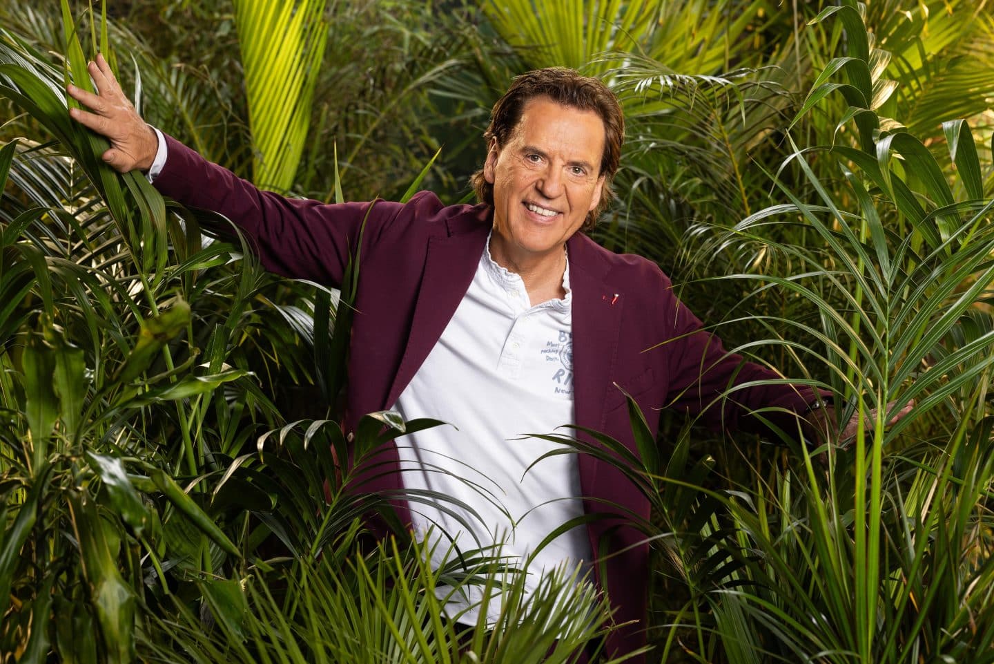 "Ich bin ein Star – Holt mich hier raus!": Wer fliegt, wer punktet? Der große Dschungelstar-Check von Jan Köppen! 12 Jörg Dahlmann, lächelnd in einem weißen Polo-Shirt und bordeauxfarbenen Blazer, mit ausgebreiteten Armen zwischen tropischen Pflanzen im Dschungel.