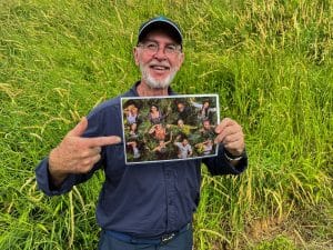 Dr. Bob, bekannt aus dem Dschungelcamp, steht vor einer grünen Wiese und hält ein Bild der diesjährigen Dschungelcamp-Kandidaten in die Kamera, während er fröhlich darauf zeigt.