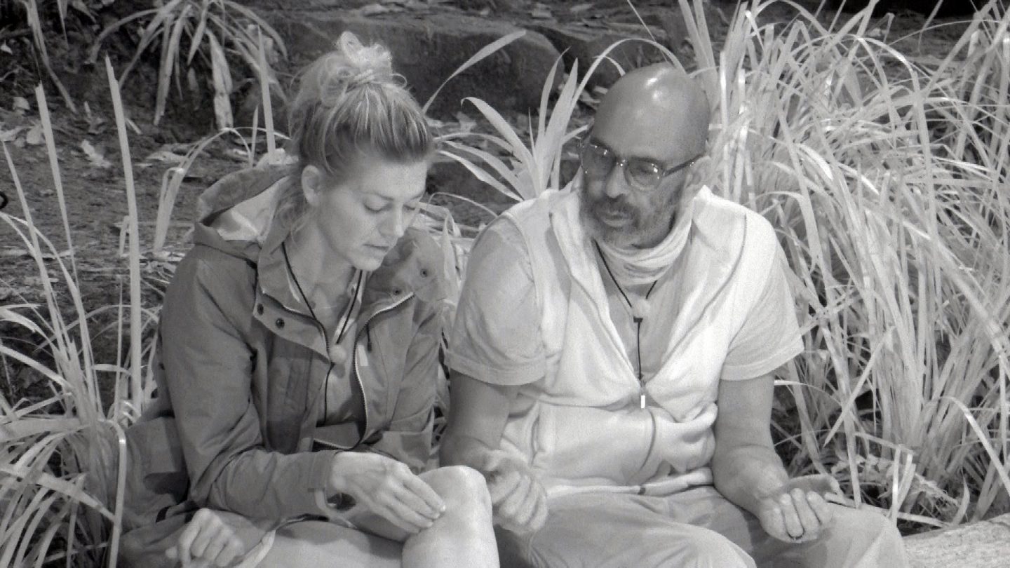 Nina Bott und Pierre Sanoussi-Bliss sitzen während der Nachtwache im Dschungelcamp auf dem Boden, umgeben von hohen Gräsern. Die Aufnahme ist in Schwarz-Weiß gehalten. Nina, mit blonden Haaren im Dutt und einer Jacke, blickt nachdenklich nach unten und berührt ihr Knie. Pierre, der eine Brille und ein helles Outfit trägt, spricht und gestikuliert mit der Hand, während er Nina ansieht. Beide wirken in ein ernstes Gespräch vertieft.