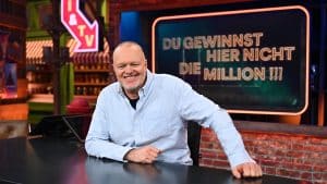 Stefan Raab sitzt lächelnd an einem Schreibtisch in einem TV-Studio. Im Hintergrund ist ein Bildschirm mit dem Schriftzug „Du gewinnst hier nicht die Million !!!“ zu sehen, der Titel seiner neuen RTL-Show.