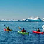 Drei Kajakfahrer paddeln durch das eisige Wasser einer arktischen Landschaft, umgeben von schwebenden Eisbergen und schneebedeckten Formationen unter einem klaren blauen Himmel.