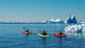 Drei Kajakfahrer paddeln durch das eisige Wasser einer arktischen Landschaft, umgeben von schwebenden Eisbergen und schneebedeckten Formationen unter einem klaren blauen Himmel.
