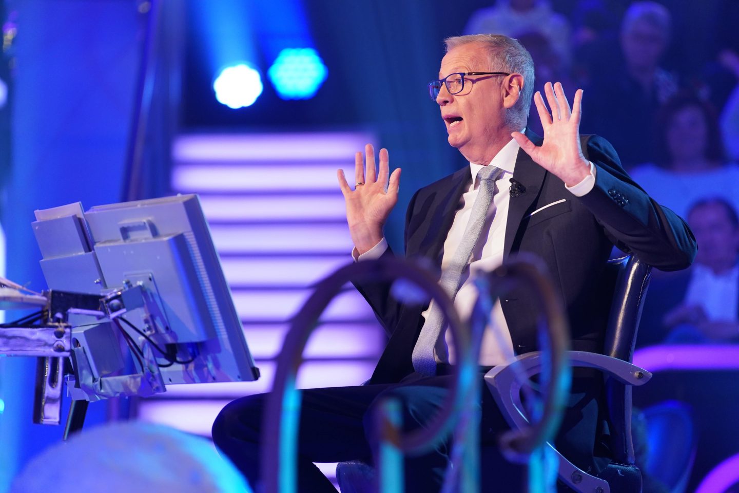 „Günther Jauch sitzt auf dem Kandidatenstuhl bei der Quizshow ‚Wer wird Millionär?‘, gestikuliert mit beiden Händen und zeigt eine lebhafte Reaktion, während er das Publikum unterhält. Im Hintergrund sind bunte Studiobeleuchtung und Zuschauer zu sehen.“