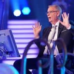 „Günther Jauch sitzt auf dem Kandidatenstuhl bei der Quizshow ‚Wer wird Millionär?‘, gestikuliert mit beiden Händen und zeigt eine lebhafte Reaktion, während er das Publikum unterhält. Im Hintergrund sind bunte Studiobeleuchtung und Zuschauer zu sehen.“