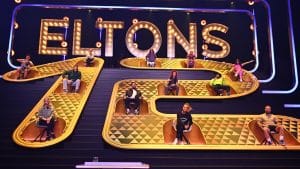 Bühne der Show „Eltons 12“ mit zwölf prominenten Teilnehmern, die in goldenen Sitzen auf einer treppenförmigen Konstruktion vor dem großen Schriftzug „ELTONS“ sitzen. Die Kandidaten sind in verschiedenen Sitzreihen positioniert und bereit für die Herausforderungen der Sendung. Die Bühne ist mit Lichtern und einem glamourösen gold-schwarzen Design gestaltet.