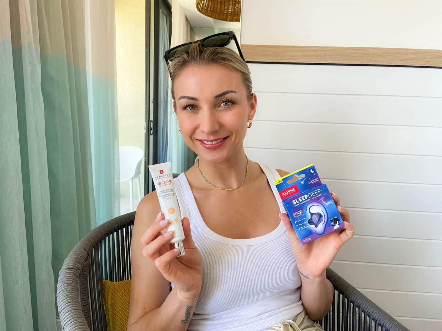 Anna-Carina Woitschack hält eine Tube BB-Creme und eine Packung Ohrstöpsel in den Händen, lächelnd in die Kamera.

