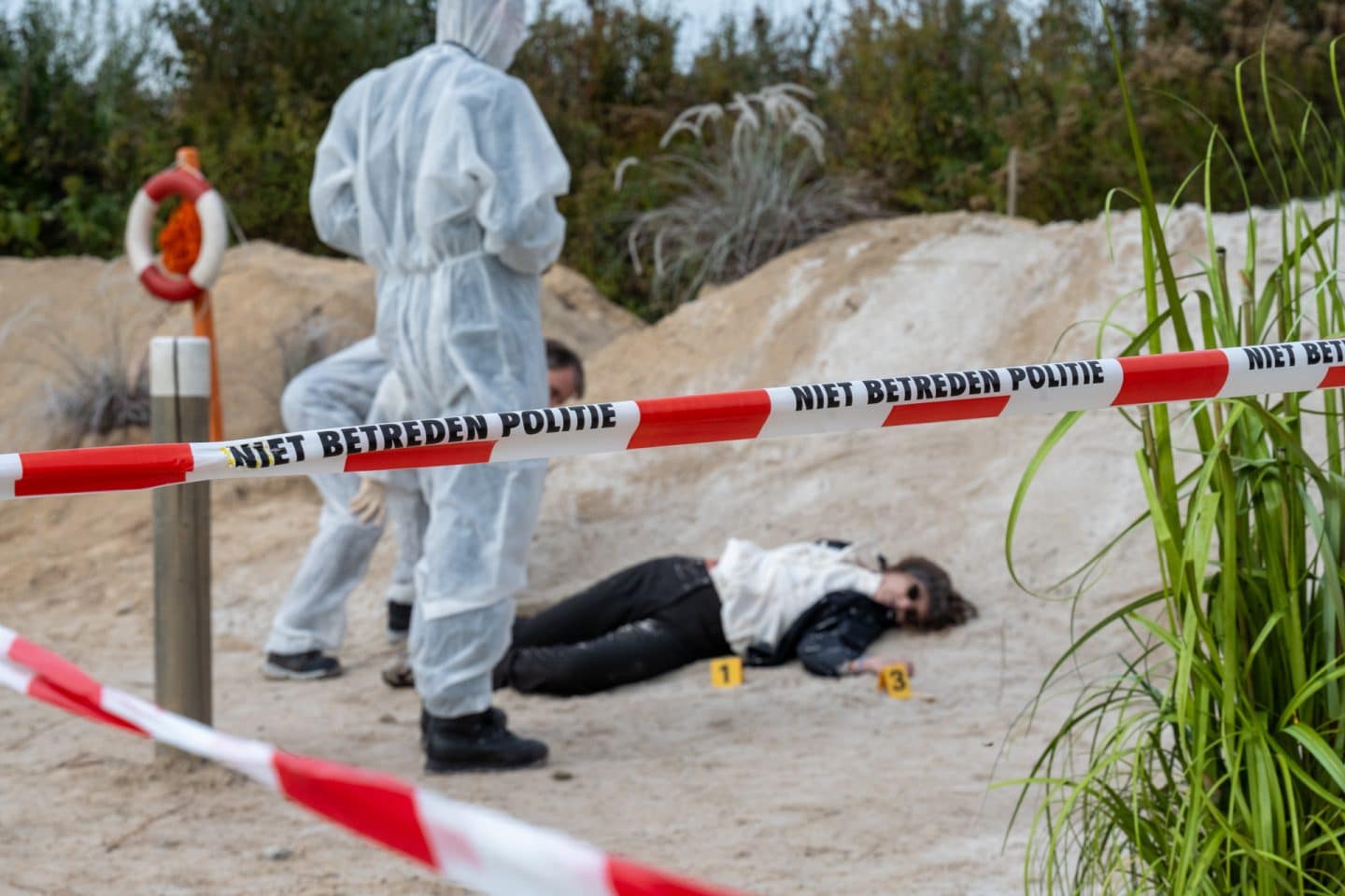 "Aktenzeichen XY... Ungelöst": Nachgestellte Szene: Rot-weiß gestreiftes Absperrband schirmt einen Tatort ab. Dahinter liegt eine tote Frau in den Dünen auf dem Boden. Neben ihr stehen zwei Personen in weißen Anzügen von der Spurensicherung und schauen sie an.